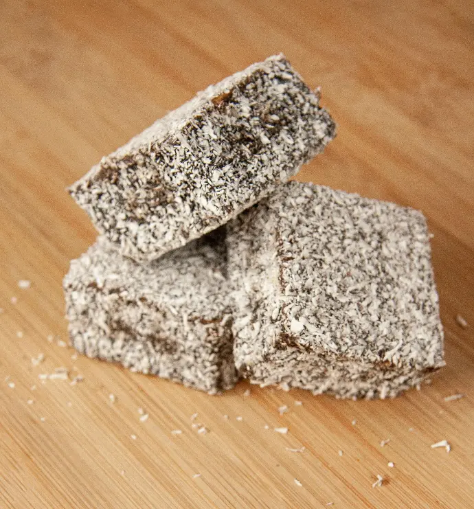 Australian lamington cakes on wooden background — chocolate and coconut treat. Close-up of three stacked chocolate-coated lamington cakes covered in shredded coconut on a wooden board.